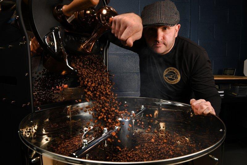 Image shows hot, freshly roasted, specialty coffee beans being dropped from black and copper coloured toper coffee roaster machine at the Flat Cap Coffee Roasting Company in Wiltshire, UK. Roaster being operated by Director wearing black top and flat cap