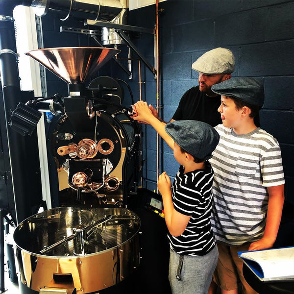 Image shows Dad teaching two young sons how to roast specialty coffee on black and copper coloured toper coffee roaster machine. Boys and Dad are wearing flat caps at the Flat Cap Coffee Roasting Company's Roastery in Wiltshire, UK